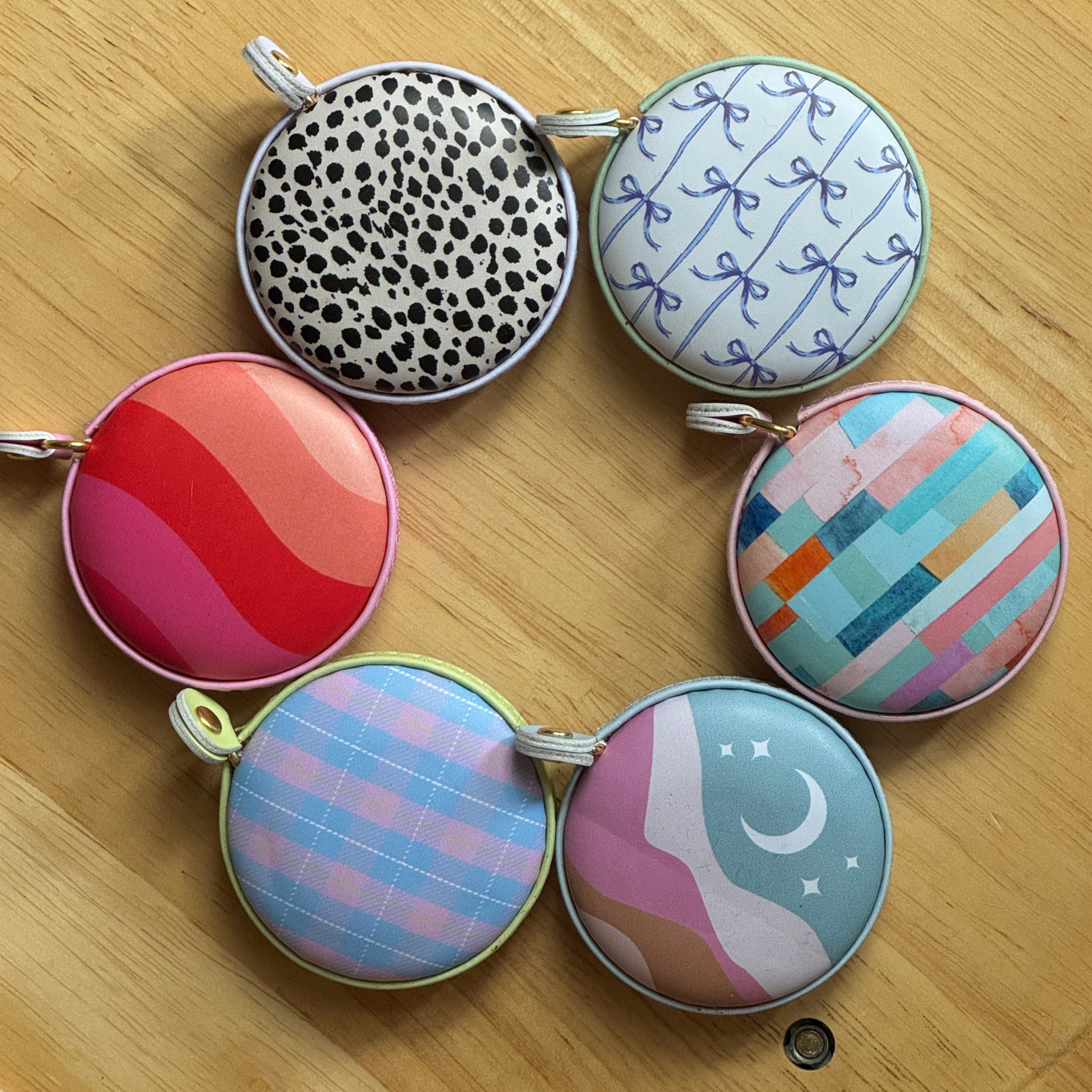 Six round fabric measuring tapes with colorful patterns and designs, arranged in a circle on a light wood surface.