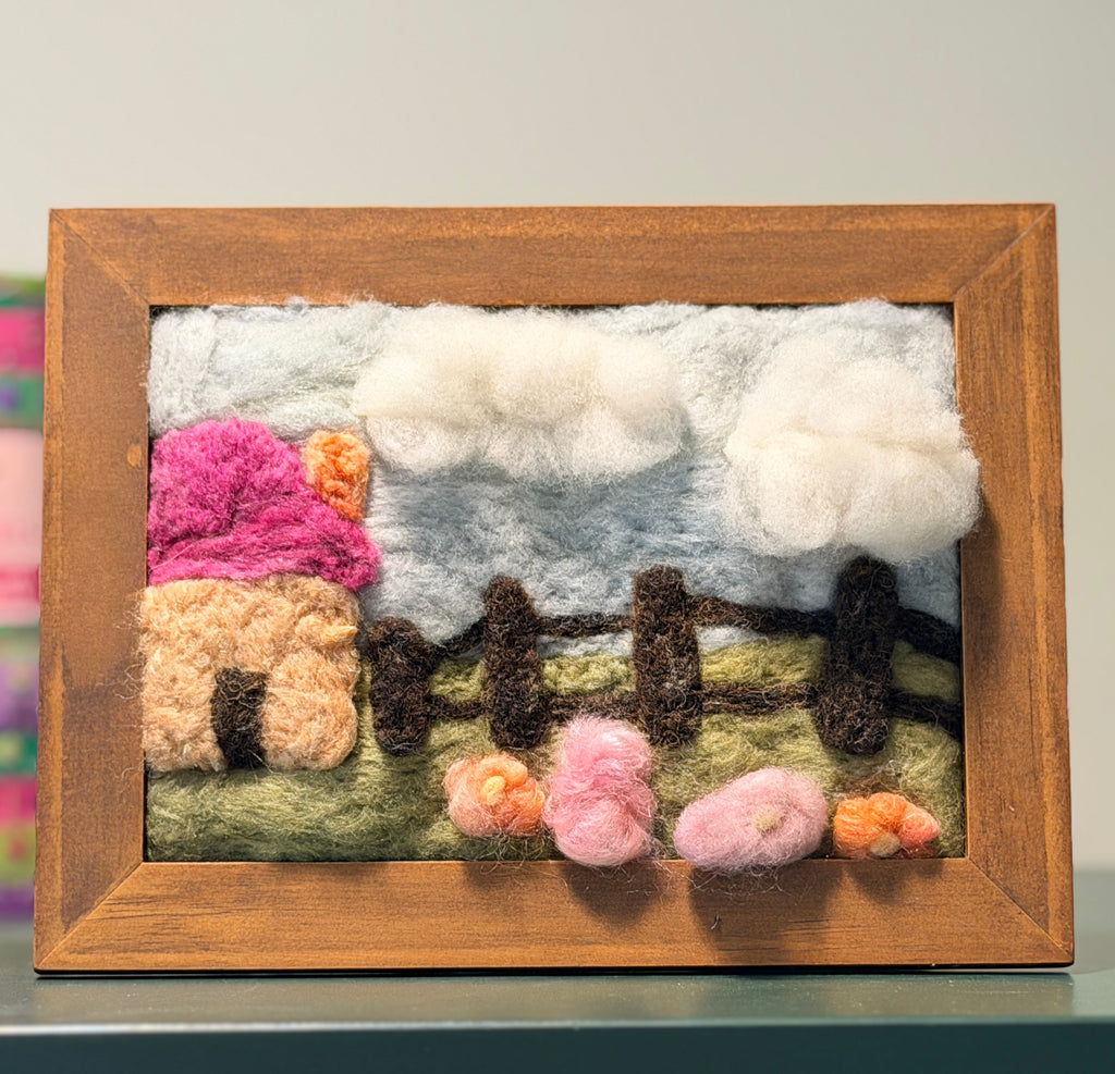 A framed wool felt landscape art piece featuring a pink and beige house, white clouds, a green field, and small flowers, set in a natural wood frame.
