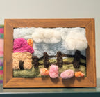 A framed wool felt landscape art piece featuring a pink and beige house, white clouds, a green field, and small flowers, set in a natural wood frame.