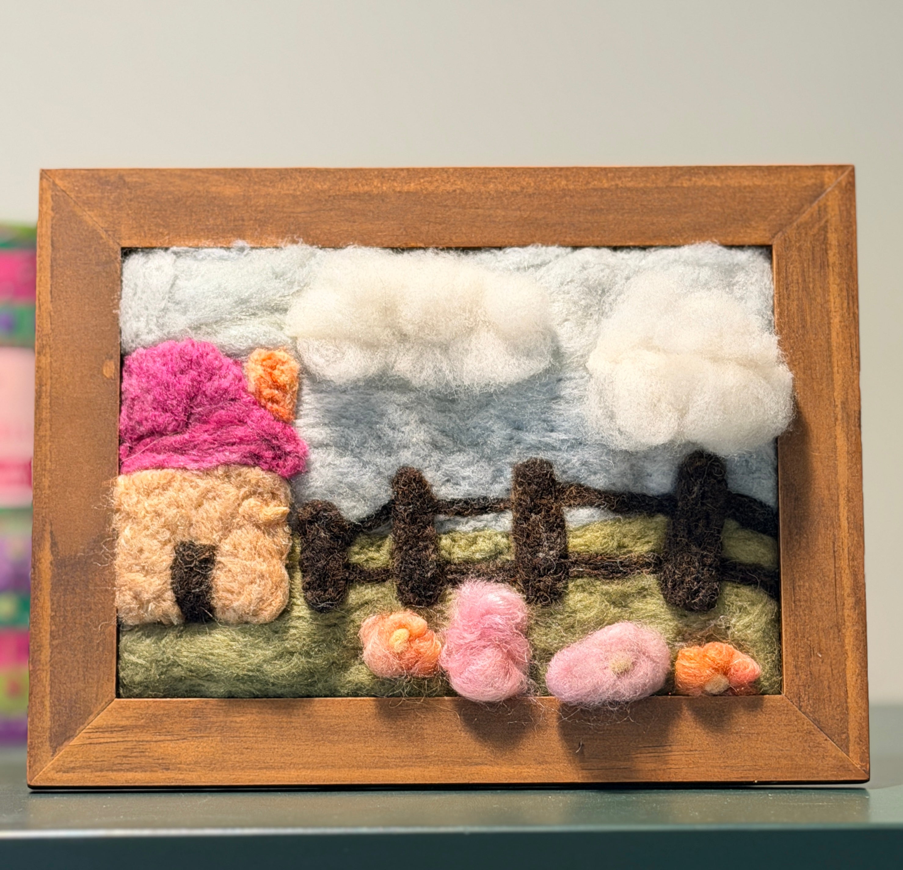 A framed wool felt landscape art piece featuring a pink and beige house, white clouds, a green field, and small flowers, set in a natural wood frame.