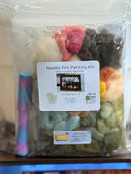 A Needle Felt Painting Kit in a clear plastic bag with a white product label. The kit contains multiple colors of wool fibers, including pink, cream, brown, green, yellow, and orange, along with a small striped paintbrush on the left.