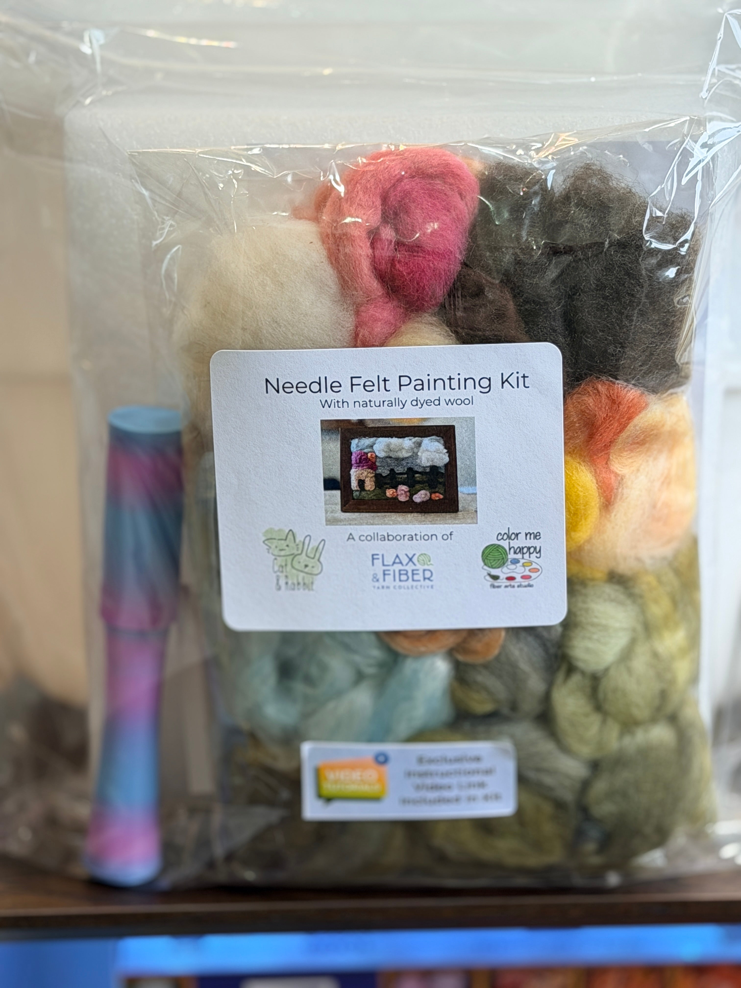 A Needle Felt Painting Kit in a clear plastic bag with a white product label. The kit contains multiple colors of wool fibers, including pink, cream, brown, green, yellow, and orange, along with a small striped paintbrush on the left.