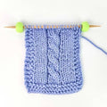 A light blue cable-knit swatch on wooden knitting needles with green ball tips, shown against a plain white background.