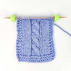 A light blue cable-knit swatch on wooden knitting needles with green ball tips, shown against a plain white background.